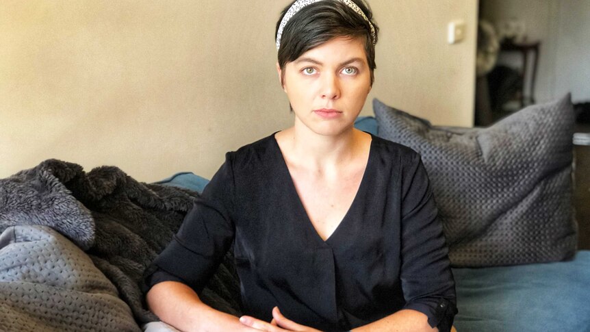 A young woman with short hair sits on her couch, looking into the camera.