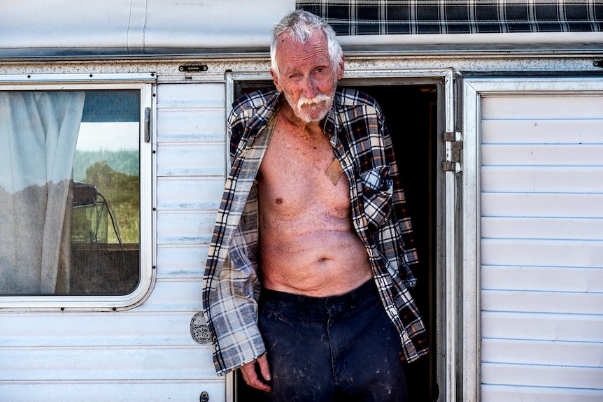 An elderly man stands in a caravan doorway.