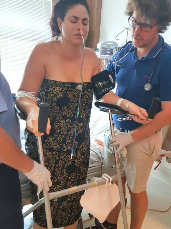 A woman on a drip with tubes in her nose tries to stand next to a hospital bed with the support of staff and crutches.