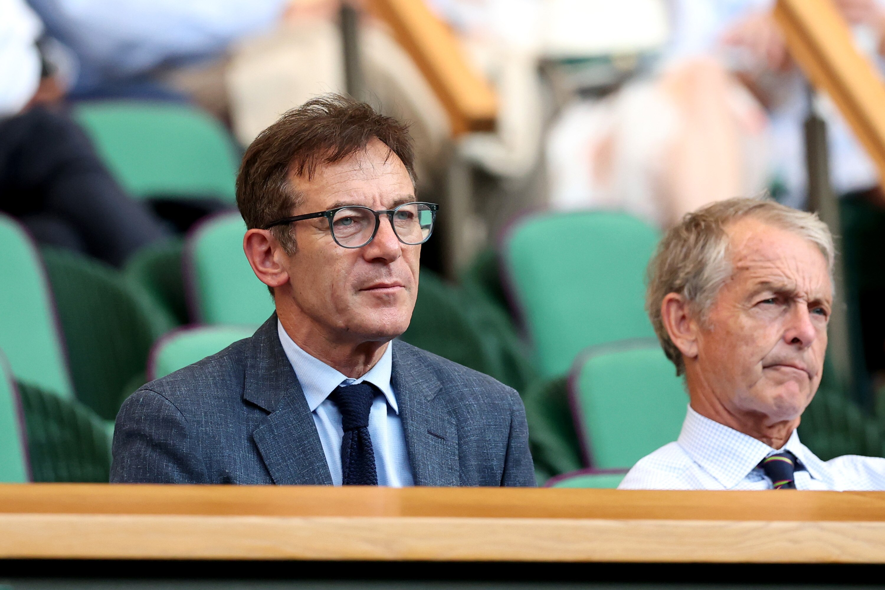 Jason Isaacs in a dark suit and glasses watches the tennis form the royal box
