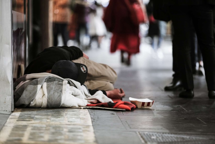 Homeless man sleeping on the footpath in a city as pedestrians walk past