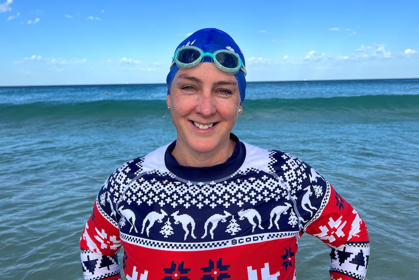 A smiling woman in swimming cap, goggles and a Christmas themed rashie stands by the surf.  