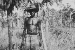monochrome of man standing in the bush wearing shorts and a hat.