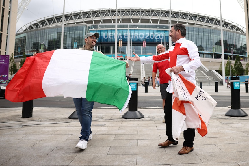 Fans stand sibe by side in front of stadium waving Italy and England flags 