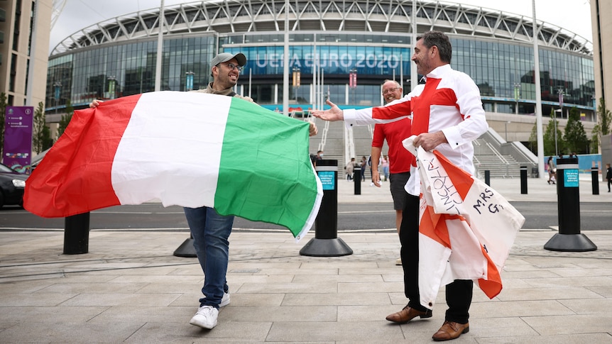 Fans stand sibe by side in front of stadium waving Italy and England flags 