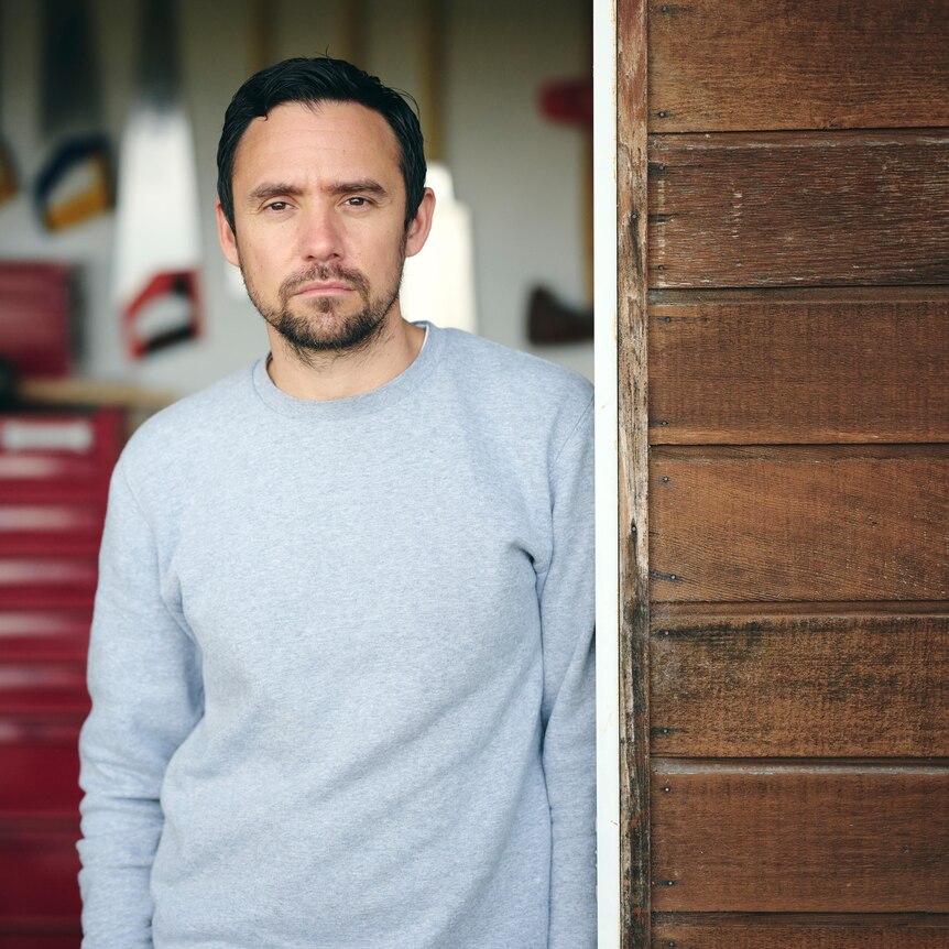 James stands in the doorway of his workshop studio wearing a grey jumper, looking meaningfully down the barrel of the camera.