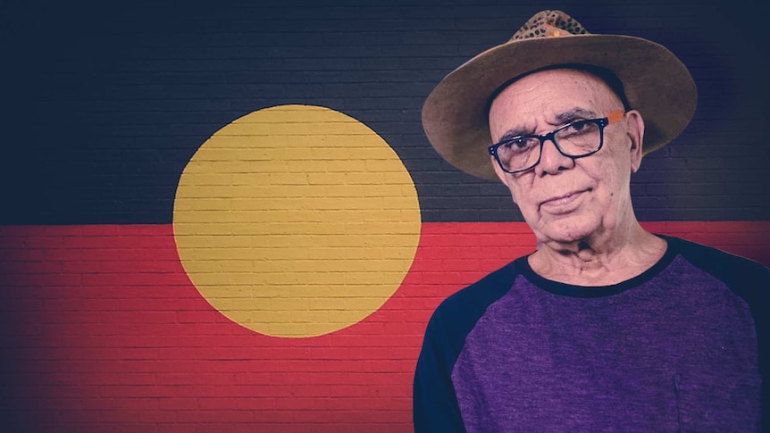 Harold Thomas stands in front of a wall painted with his Aboriginal Flag design.