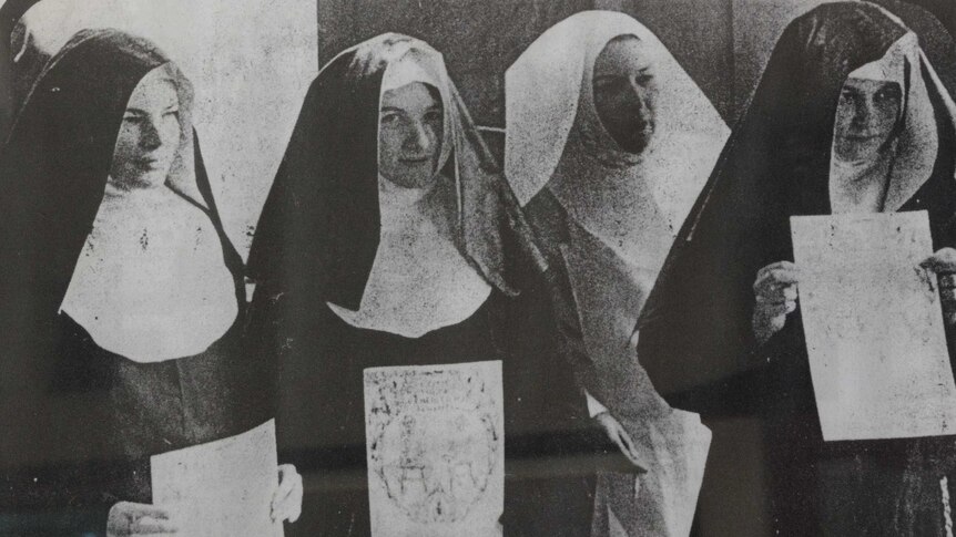 Four women in nuns costumes line up holding A4 signs, the writing on them is unclear.