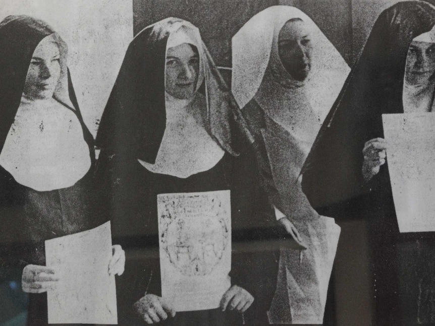 Four women in nuns costumes line up holding A4 signs, the writing on them is unclear.