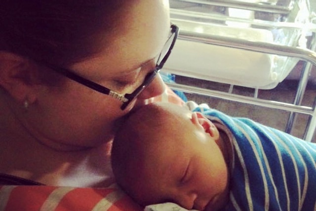 A woman sits holding a newborn boy and kissing him on the head.