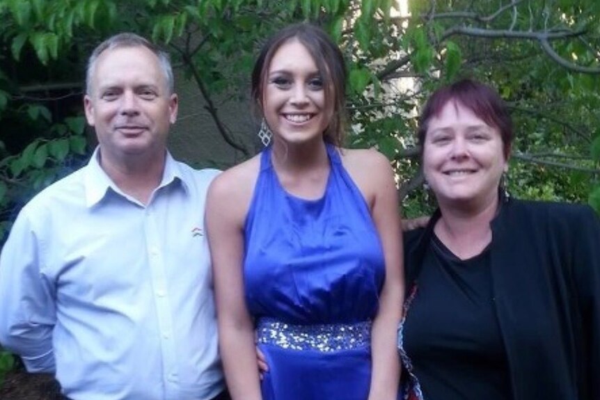 A young woman in a formal dress stands smiling with her parents.