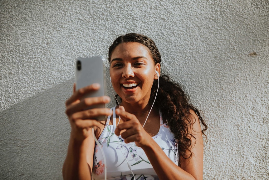 A girl wearing exercise gear and earbuds smiles at a mobile phone in her hand.