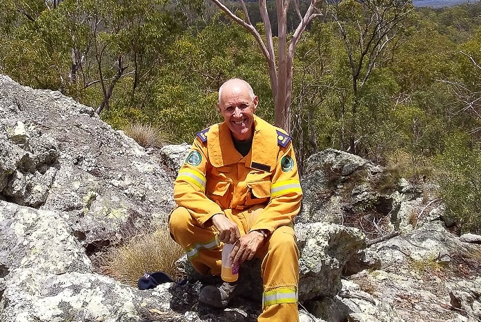 A man in a yellow fire brigade uniform sits on top of a rock surrounded by bushland