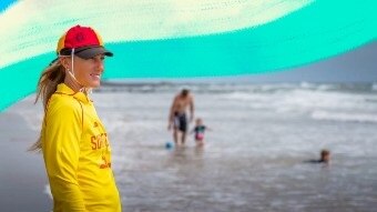 Surf lifesaver on the beach in her uniform looking out towards the ocean, with a father and child in the background.