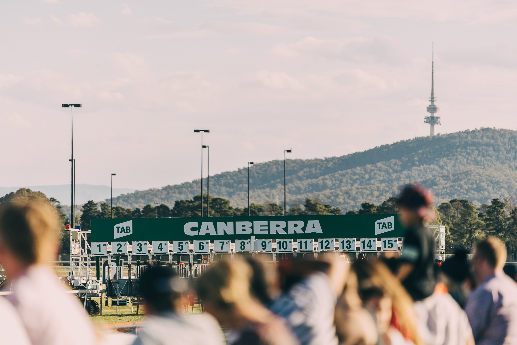 People look at the gates at a horse-racing track; in the background is a mountain with a large tower on top.