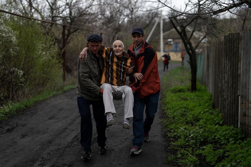 Two men carry an elderly man down a driveway