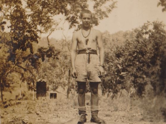 Sepia image of young man in army boots, shorts, no shirt and crewcut standing above a trench and surrounded by trees