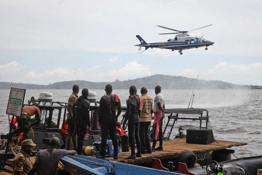 A group of six men standing on a platform look up at a helicopter hovering low above water.