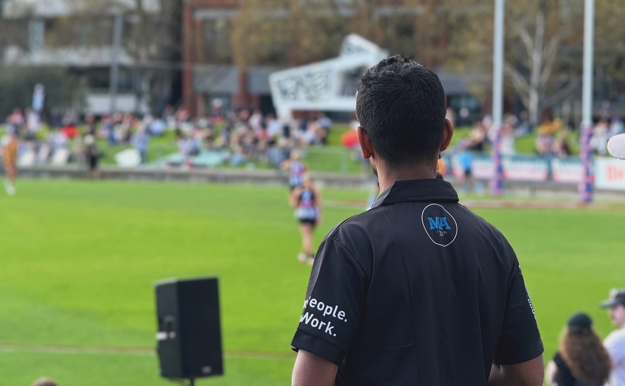 Una foto tomada detrás de un hombre de cabello oscuro con una camiseta negra con el logo de MA viendo un partido de fútbol.