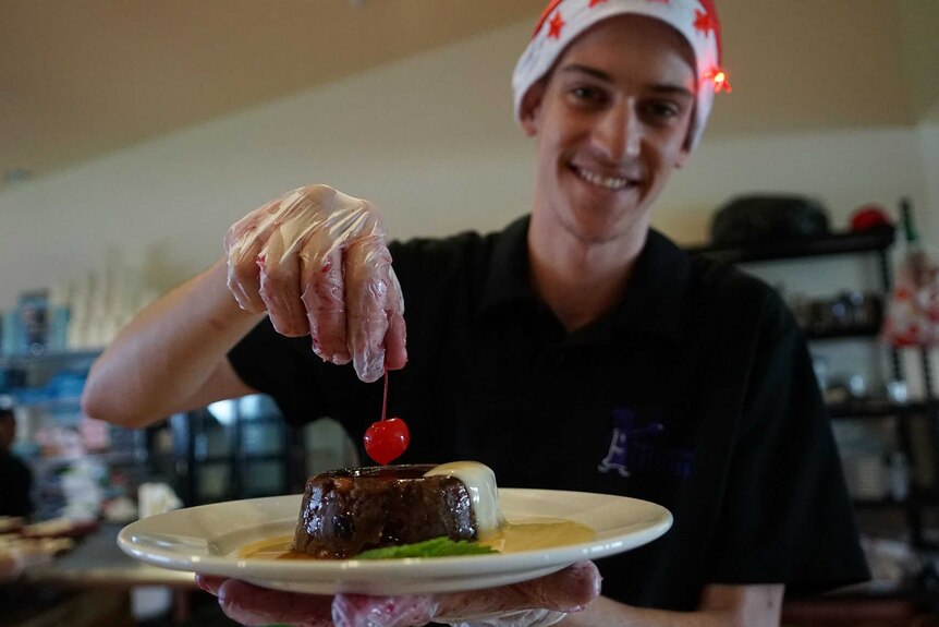 Benny Dawson puts the cherry onto the Kakadu plum pudding and smiles at the camera. He is wearing a Santa hat and black shirt.