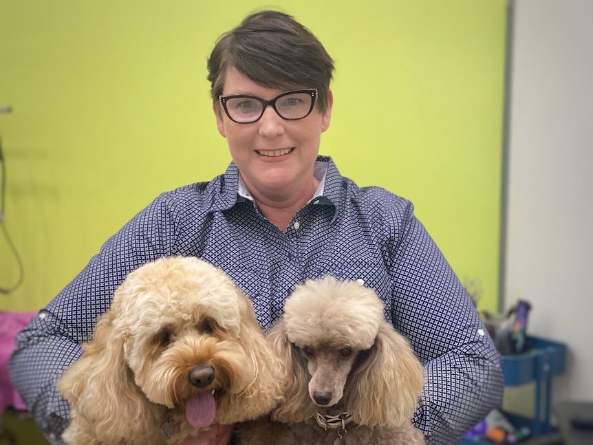 A woman with short hair and glasses smiles with two dogs sitting on her lap