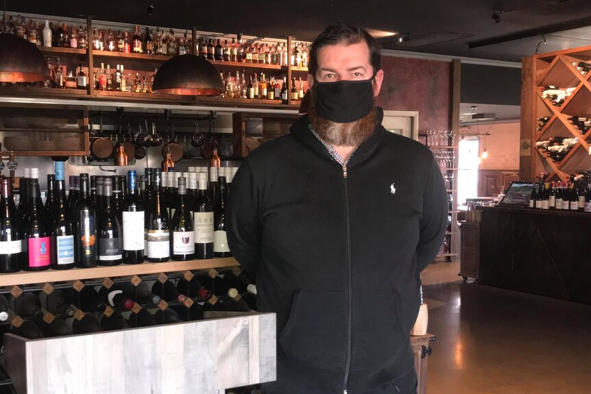 Hombre con una máscara negra de pie delante de un estante de vino