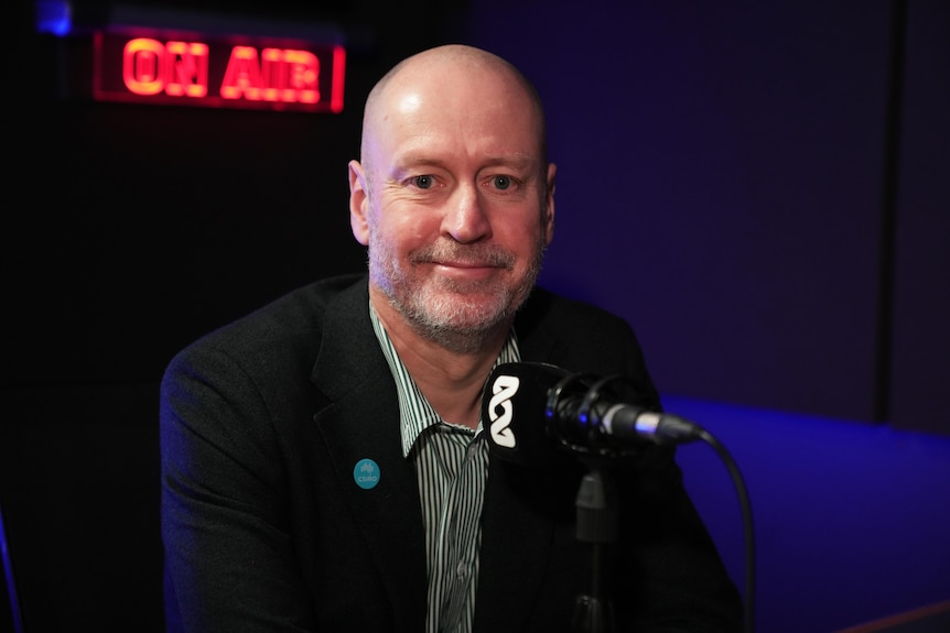 A bald man sitting at a ABC branded microphone with an on air sign lit up in the background. 