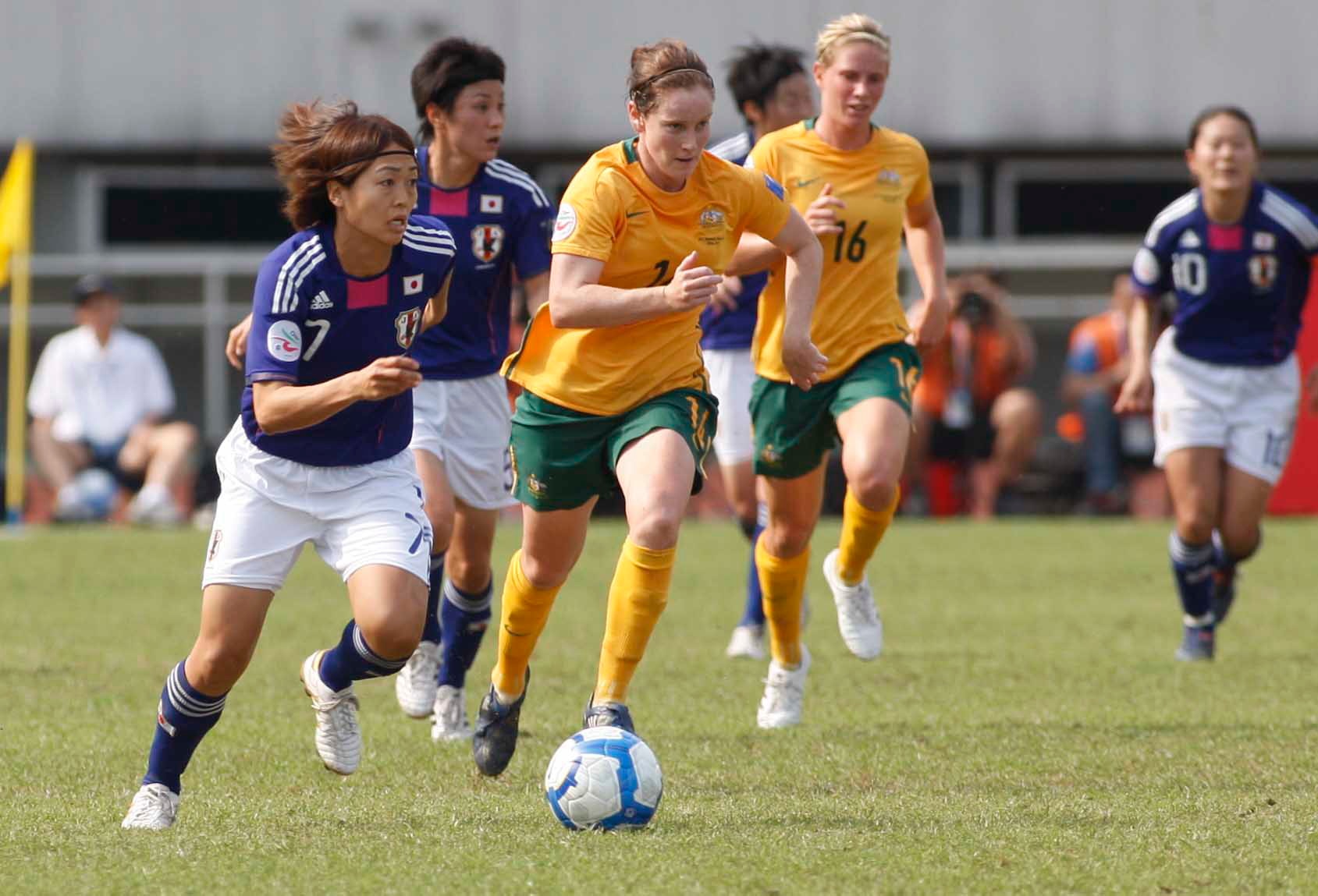 Um jogador de futebol de azul corre com a bola, sendo perseguido por um jogador de verde e dourado