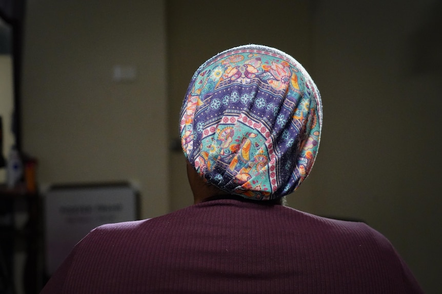 The back of a woman's head, covered in a colourful scarf, as she sits inside a dark room.