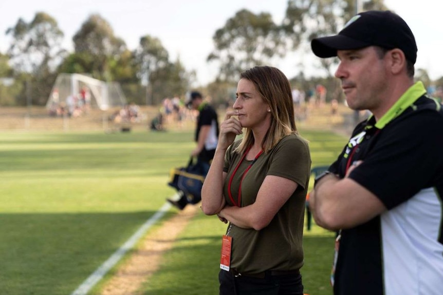 A women's soccer coach stands pitch-side with her hand in front of her mouth, watching a match.