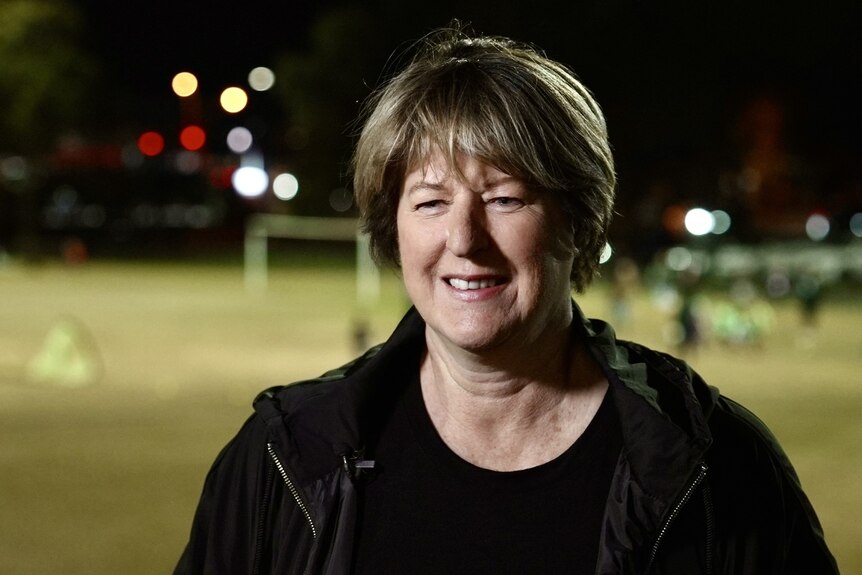A middle-aged white woman with short brown hair. She is in front of a suburban football pitch at night, lit by flood lights