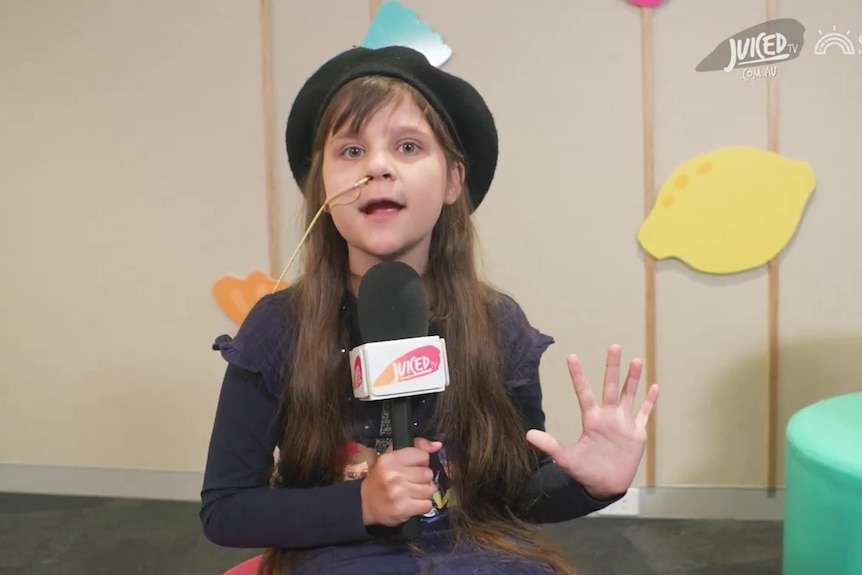 A young girl wearing a beret talks to the camera while holding a microphone in a studio.