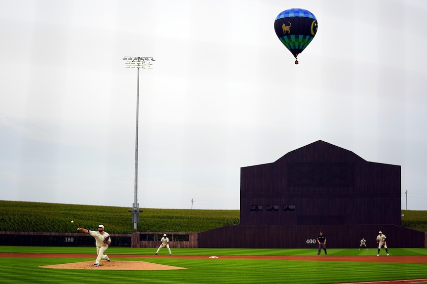 A hot air balloon is seen over a field, where a pitcher is throwing the ball