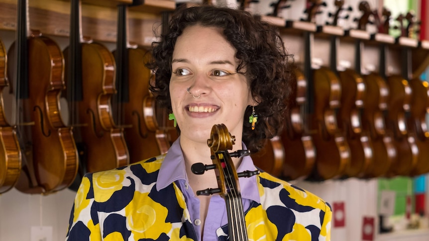 Sophia Mackson holds a viola vertically in front of her body. She stands in front of a wall of violins.