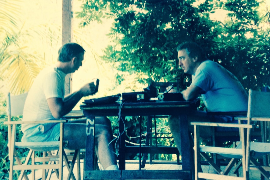 Doi bărbați așezați la o masă pe un balcon.  Bărbatul din stânga ține o ceașcă, cel din dreapta se sprijină pe masă.