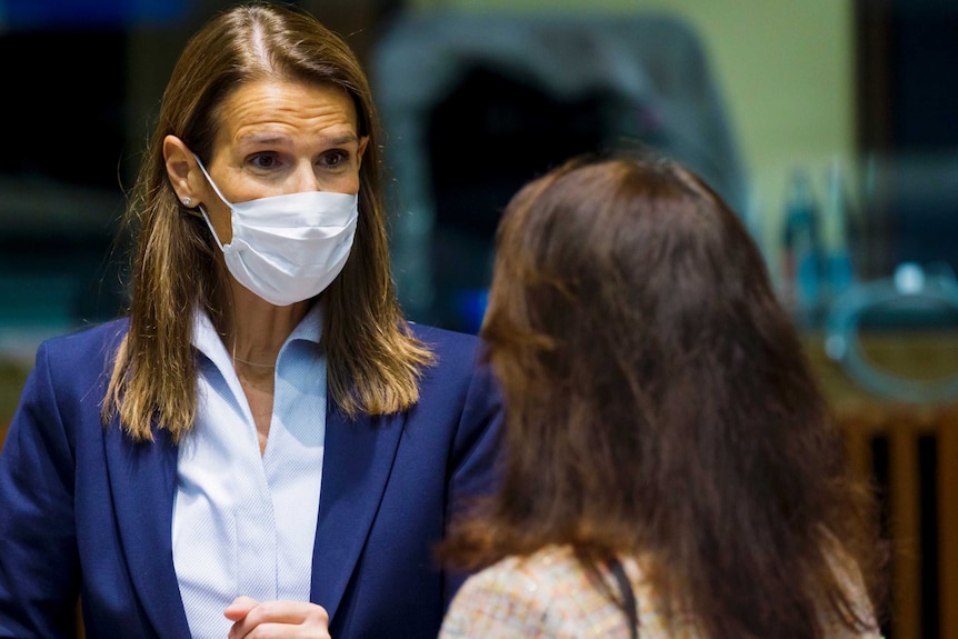 A woman wearing a suit and a face mask faces the camera while speaking to another woman.