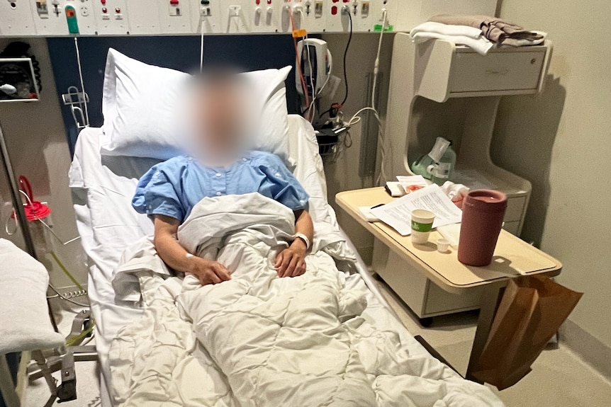 A man in a blue hospital gown, lying in a hospital bed with his face blurred.