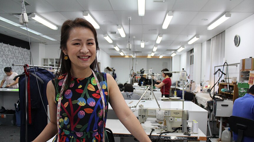 A woman with a measuring tape draped around her neck stands behind a table with sewing machines and workers in the background.