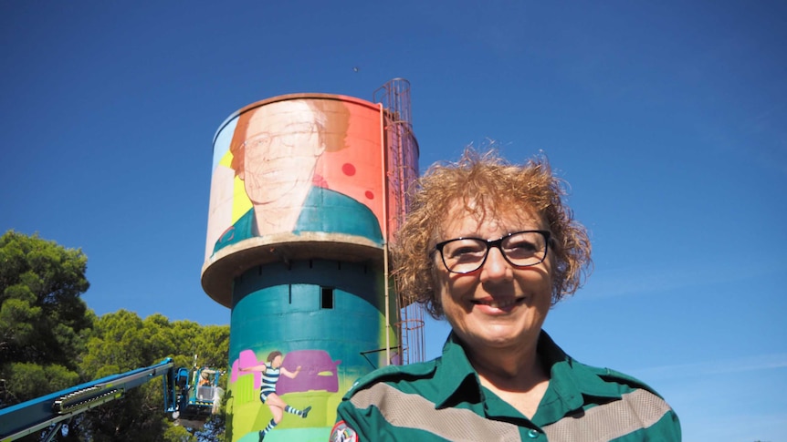 Lady stands in front of colourful water tower in green SA Ambulance uniform which depicts her face
