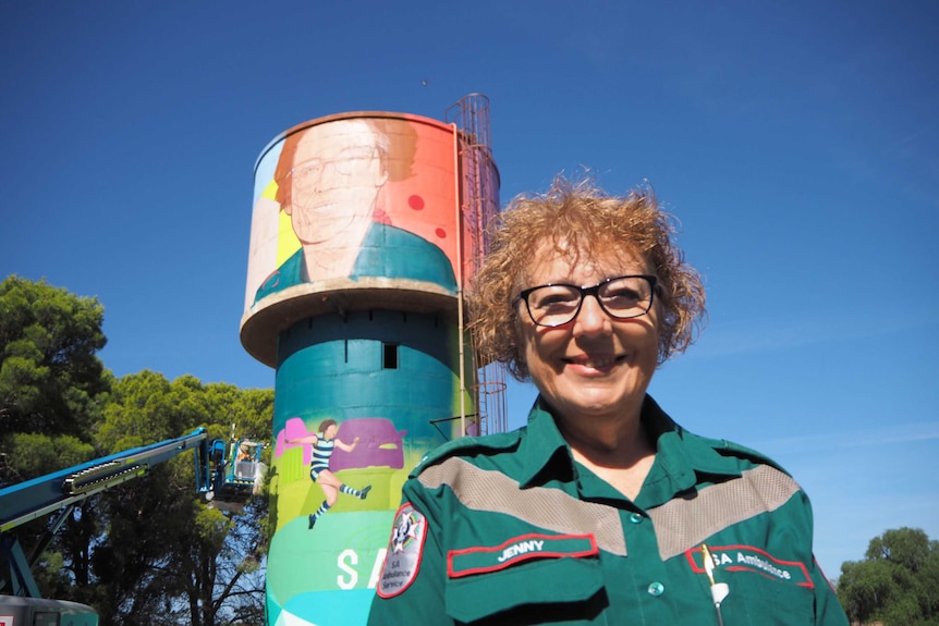 Lady stands in front of colourful water tower in green SA Ambulance uniform which depicts her face