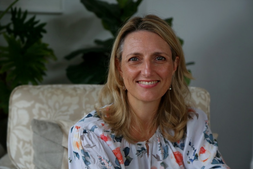 A portrait of a middle-aged woman with blonde hair on a sofa seat in her home. 