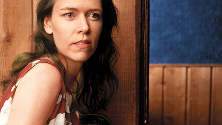 Gillian Welch sits in a wood-panelled room, wearing a dark red dress.