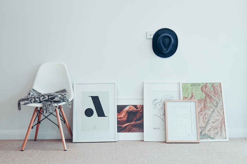 Framed photographs and artwork sit on the carpet beside a chair, ready to be hang on the wall as a DIY project.