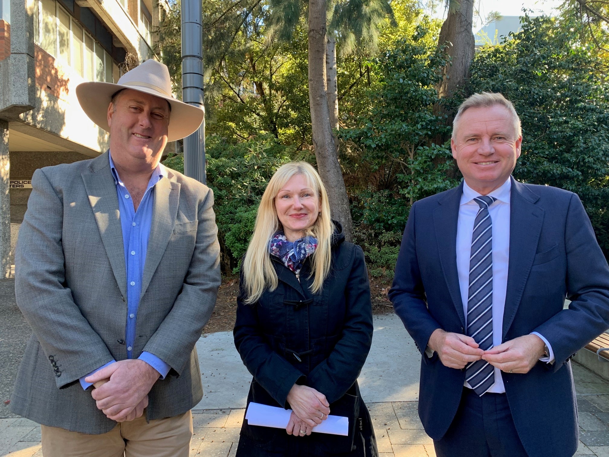 Two men, one wearing a grey suit and a hat, the other a blue suit, flank a shorter blonde woman.