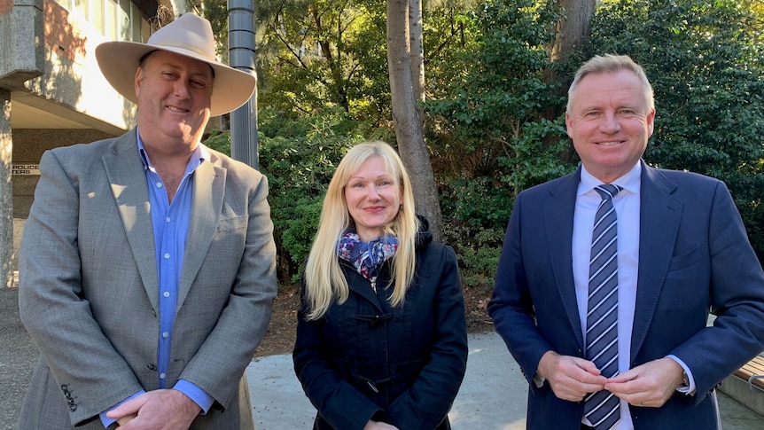 Two men, one wearing a grey suit and a hat, the other a blue suit, flank a shorter blonde woman.