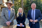 Two men, one wearing a grey suit and a hat, the other a blue suit, flank a shorter blonde woman.