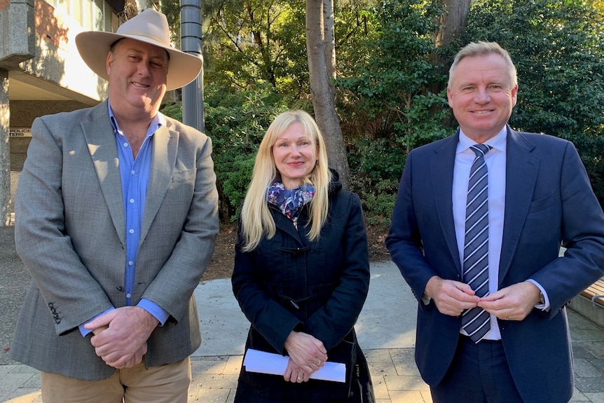 Two men, one wearing a grey suit and a hat, the other a blue suit, flank a shorter blonde woman.