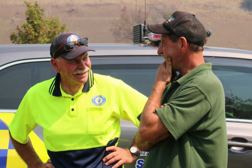 Two men wearing caps and polo shirts stand by a car and chat.