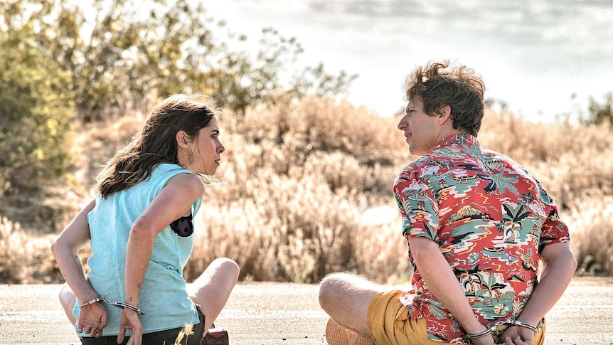 Cristin Milioti and Andy Samberg sitting in the desert handcuffed in a scene from Palm Springs