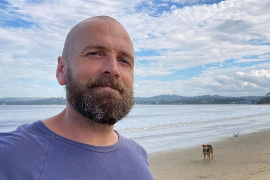 A man with short cropped hair and a short beard, wearing a T-shirt, on a beach with the ocean behind him.